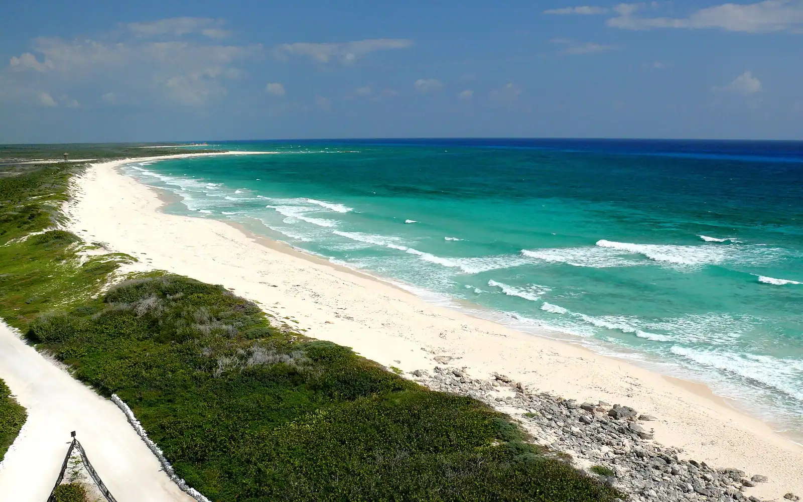 A slim white lighthouse tower rising above low coastal scrub at the southern tip of a Caribbean island