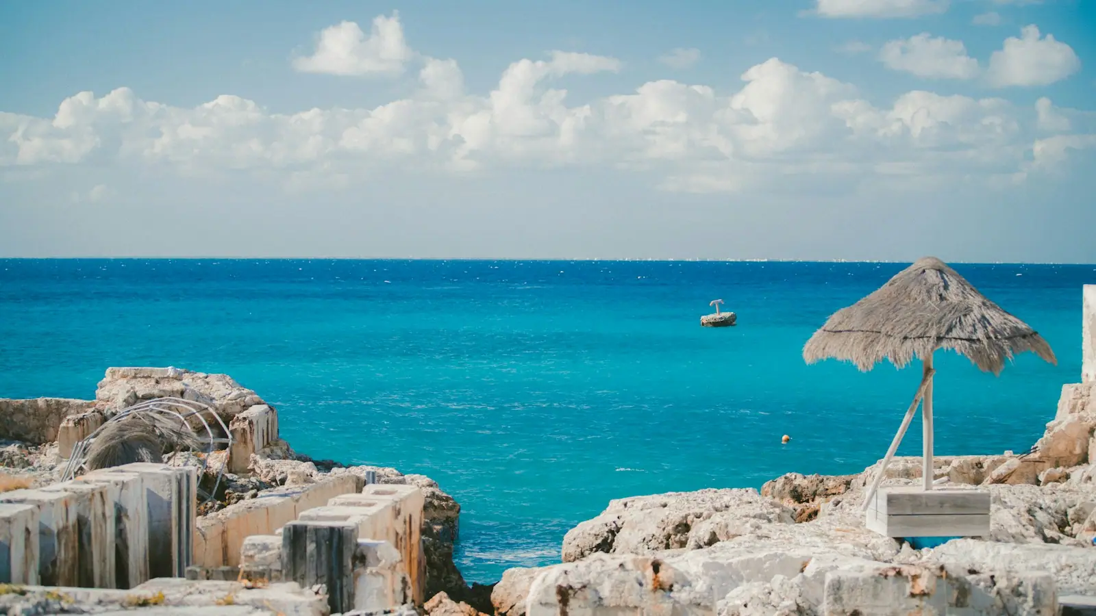 A thatched shade shelter beside low stone ruins on a coastal site with the Caribbean behind