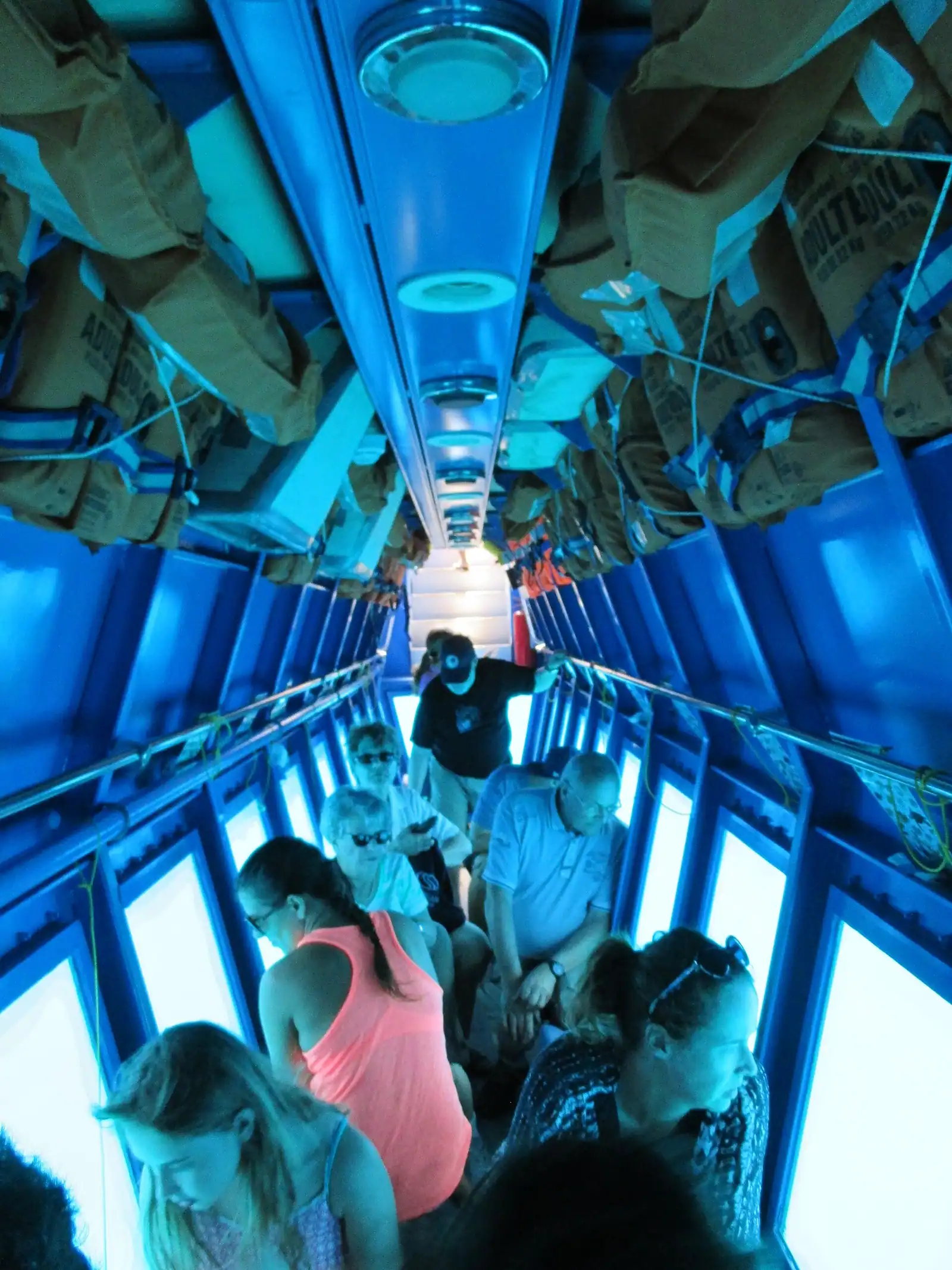 Passengers seated along the cabin of a tourist submarine, each at a round porthole looking out into blue water